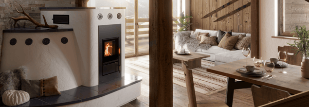 Ein weißer Kaminofen steht in einem rustikal eingerichteten Wohnbereich gegenüber einer Couch und neben einem Esstisch aus Holz.