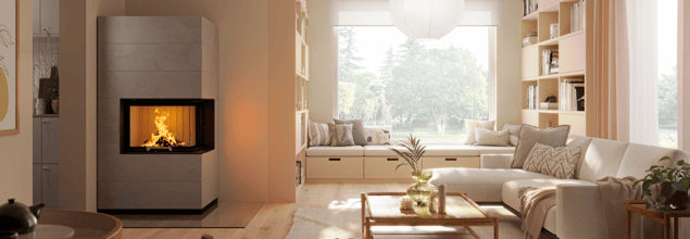 Eine graue Kaminanlage steht in der Ecke eines hell eingerichteten Wohnzimmers neben einem Sofa und einer Sitzecke mit großem Fenster.