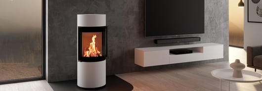 Der Kaminofen Passo XS steht neben einem an der Wand hängenden Fernseher in einem minimalistischen Wohnzimmer.