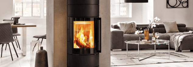 Ein schwarzer Kaminofen steht vor einer schmalen Betonwand, die als Raumteiler zum Wohnzimmer dient.