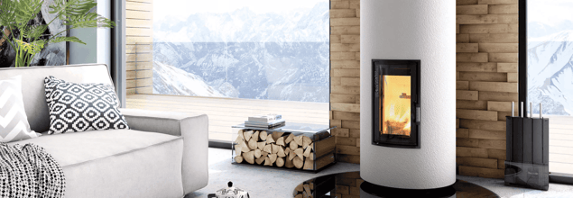 Ein weißer Kaminofen  mit einem Holzfach daneben steht vor einer gemauerten Wand zwischen zwei großen Fenstern in einem modernen Wohnzimmer.