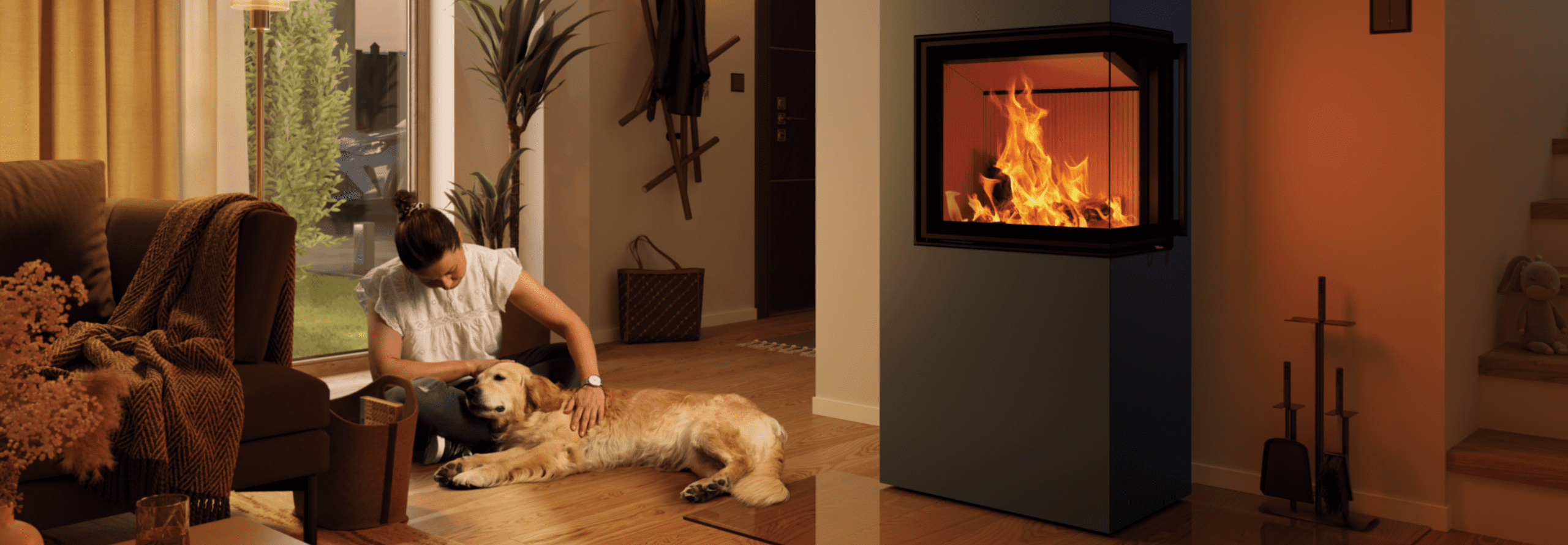 Ein Hund liegt auf dem Boden eines Wohnzimmers neben einem Kaminofen und einer Couch und wird von einer Frau gestreichelt.