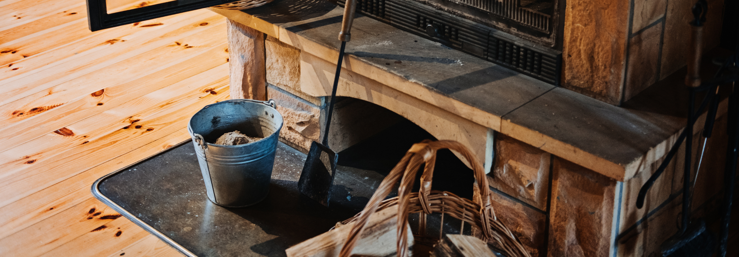 Vor einem Kaminofen steht ein Ascheeimer und eine Schaufel zur Kaminreinigung.